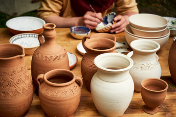 A man in old medieval Byzantine clothes is sitting painting clay dishes with a brush. Pottery in nature in retro style.