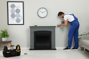 Professional technician sealing electric fireplace with caulk near white wall in room