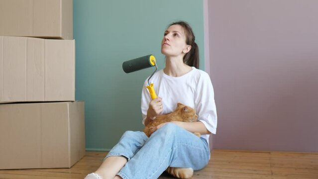 Young beautiful woman with a cat in her arms in a new house around cardboard boxes, looks around and plans how to disperse the furniture. An independent strong woman. Moving to a new house