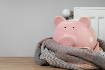 Piggy bank wrapped in scarf on wooden table near heating radiator, space for text