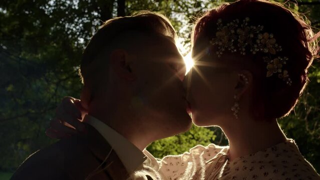 bride and groom are standing in forest in sun, setting sun is shining into camera, beautiful outline on people. Beautiful loving couple of man and heterosexual woman kiss in sun in park