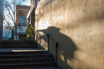 A woman is walking up a wide concrete outdoors stairs. The sun is off to her side and creating a large shadow on the wall next to her. A two story building is in the background.