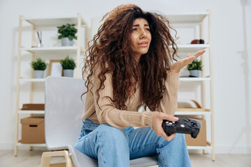 Upset frustrated tanned pretty curly Latin lady lose in game with joystick gamepad sit on chair in home modern interior look up aside. Copy space Mockup Banner. Concept gamer, emotions