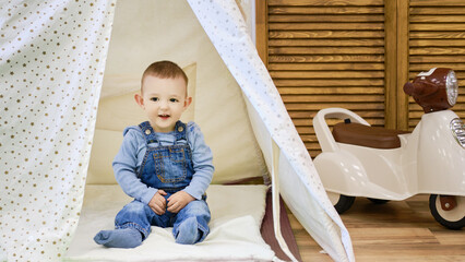 Happy toddler baby boy sitting in a children tent, playroom. Kid age one year