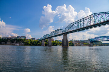 Fototapeta na wymiar the Walnut Street Bridge over the rippling blue waters of the Tennessee river with rocks along the banks surrounded by lush green trees and grass with blue sky and clouds at Coolidge park