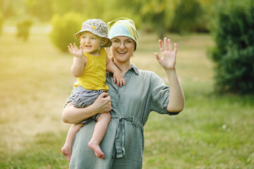 Mother holds a happy toddler baby boy in her arms and waves her hand. Smiling child with mom doing hello or bye gesture. Kid age one year