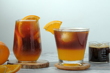cold Iced black coffee mixed with orange juice and orange slices in glass on wooden plate in white background with nature leaves silhouette shadow. two tones layer fresh summer drink in the morning