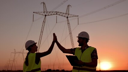 Two electrical engineers give high five against the backdrop of high-voltage electrical tower at sunset. Meeting of workers near power grids. Teamwork, signing contract, two businessmen