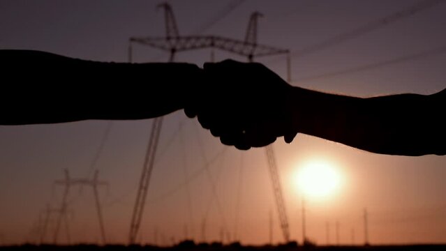 Handshake of two electrical engineers against the backdrop of high-voltage electrical tower at sunset. Silhouette of hands close up. Teamwork, signing contract, agreement between two businessmen