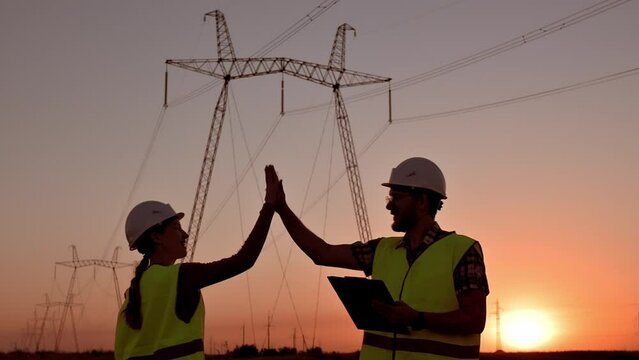 Two electrical engineers give high five against the backdrop of high-voltage electrical tower at sunset. Meeting of workers near power grids. Teamwork, signing contract, two businessmen