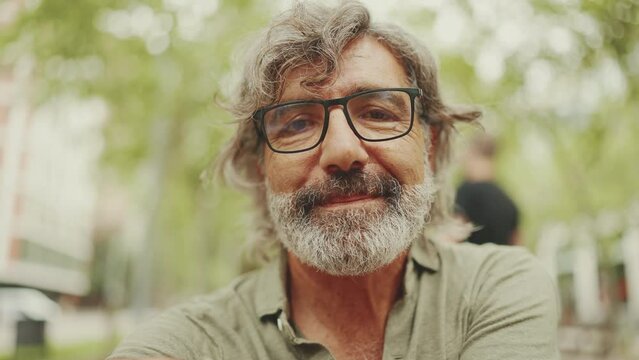 Clouse-up, thoughtful middle-aged man with gray hair and beard wearing casual clothes sits on bench. Mature gentleman in eyeglasses turns his head and looks into the camera and smiles