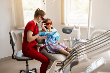 Child prepare for Uv illumination of photopolymer tooth filling in dentistry. Dentist in red...