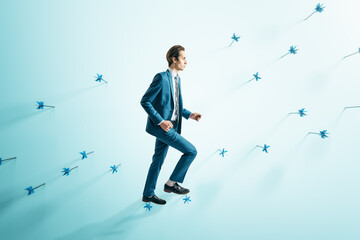 Attractive businessman in suit walking on abstract wall with many dart arrows. Goal setting and...