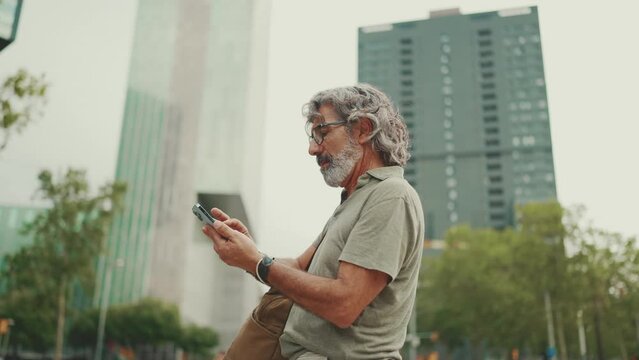 Friendly middle aged man with gray hair and beard looking at map trying to find his way using his mobile phone. Mature gentleman in eyeglasses using map app in cellphone outdoors