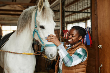 Portrait of young African American woman stroking white horse in stables and smiling happily