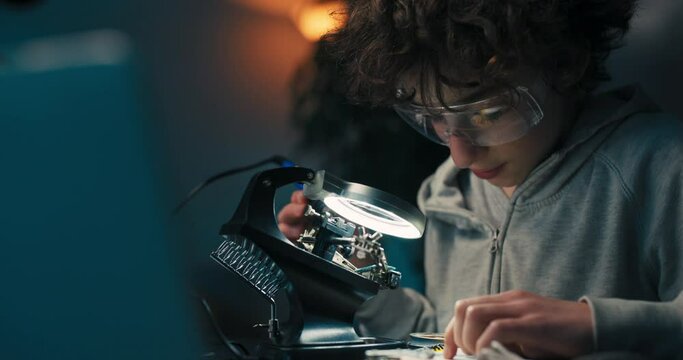 Afro boy wearing safety glasses soldering motherboard in room at desk. The child is developing a hobby for electronics and is working on a school project.