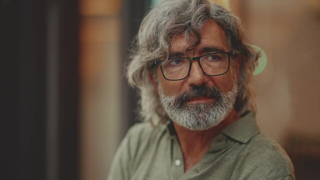 Clouse-up, thoughtful middle-aged man with gray hair and beard, wearing casual clothes, sits in street cafe. Mature gentleman in eyeglasses drinks aroma coffee