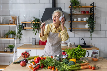 Senior attractive woman listening to music and singing in her headphones in the kitchen. Active...