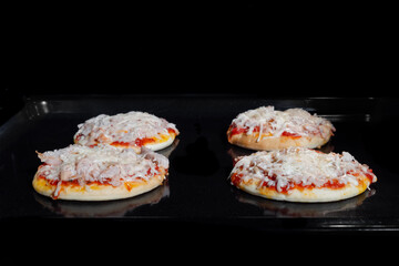 Four baked mini pizzas with ham, sause, cheese on tray in electric oven - front view, black background. Italian cuisine, homemade bakery, fast food, cooking concept