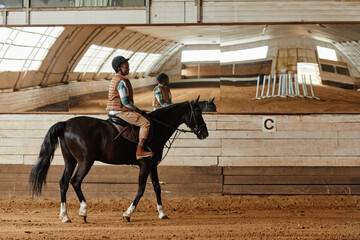 Full length side view at young woman riding horse in indoor arena or practice stadium with mirrors, copy space