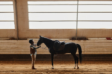 Wide angle side view portrait of young woman stroking black horse in indoor riding arena during...