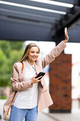 Attractive smiling woman is raising arm to say hello. She is wearing trendy hat and holding smartphone in the street