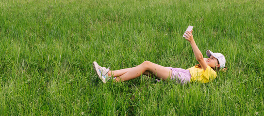 A preteen girl lies on a green grass lawn and takes a selfie on a mobile phone. Horizontal frame