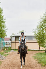 Vertical full length portrait of young black woman riding horse towards camera in outdoor ranch