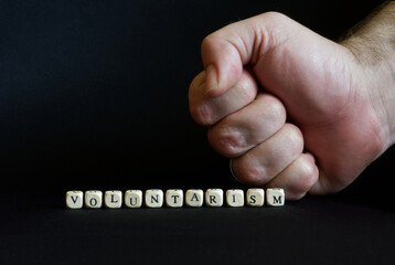 Inscription voluntarism and a clenched male fist on a black background. The concept of voluntaristic oppression, coercion and lack of freedom.