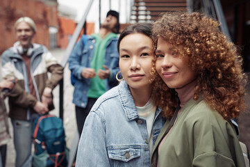 Two happy young women in stylish casualwear looking at camera while standing against teenage guys having chat by staircase outdoors