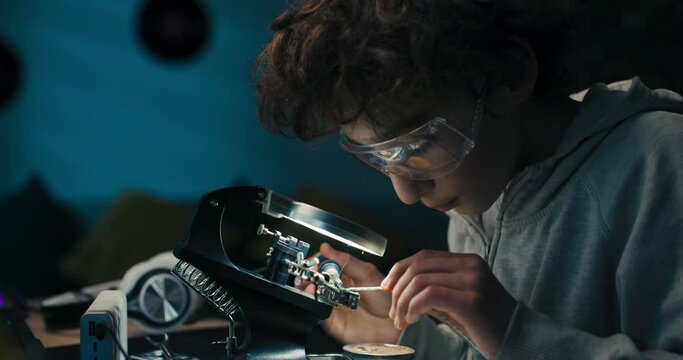 Little boy with safety glasses, future electronics technician, learning how to solder microprocessor cables and printed circuits, looking through a magnifying glass, repairing and testing electronics.