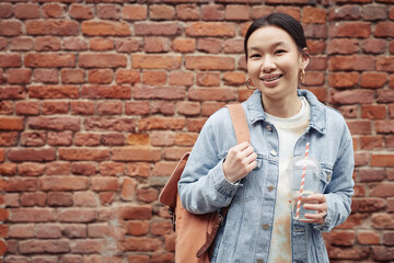 Happy Asian teenage girl in blue denim jacket and white t-shirt holding soda and backpack while standing by brick wall and looking at you