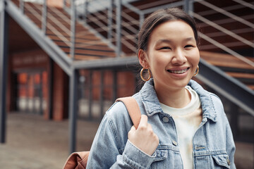 Young cheerful woman of Asian ethnicity looking at camera with toothy smile while passing by brick building with staircase
