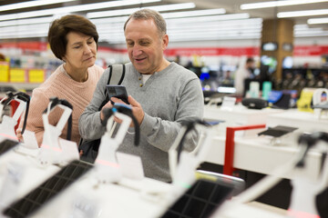 European man who came to an electronics store with his wife chooses a mobile phone to buy it