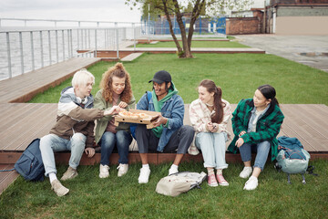 Group of intercultural teenage friends sitting on wooden pier by green lawn and having pizza while black guy holding square box