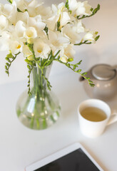 Vertical closeup of white freesia flowers in glass vase with digital tablet and tea cup and pot in background (selective focus)