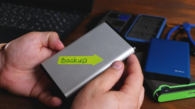 Freelancer's hand take SSD solid-state disk with back up text on laptop background. External hard drive. Save information, data storage. Plug and play device