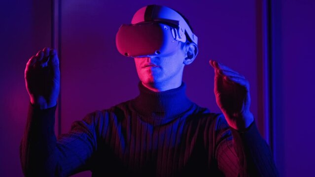 Young man in VR helmet interacts with virtual reality using his hands. Male play games, see 360 media content in VR headset under red-blue light. Futuristic technology. Immersion in metaverse concept