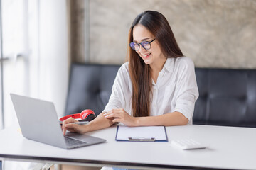 Image of young beautiful joyful asian businesswoman or asian student smiling in eyeglasses while working with laptop in home office