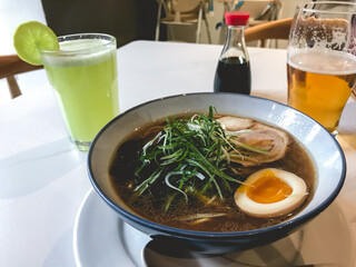 Big traditional bowl of 'ramen' (japanese noodle dish with broth, soy sauce, miso, egg, sliced pork and green onion), glass of beer and mint lemonade in a table with white tablecloth