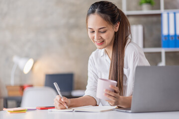 Beautiful young Asian woman working on a laptop computer while sitting in the office room, drinking coffee