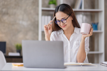 Beautiful young Asian woman working on a laptop computer while sitting in the office room, drinking coffee