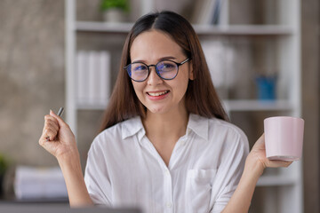 Beautiful young Asian woman working on a laptop computer while sitting in the office room, drinking coffee