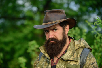 Fototapeta premium Portrait of adventure serious man extreme explorer. Man hunter in camouflage outdoor. Brutal hunter, bearded man in the wild forest in the autumn. Portrait man in cowboy hat.