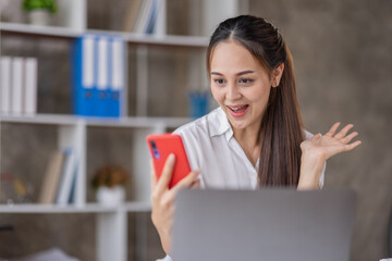 Fototapeta premium Happy Asian young woman waving hands looking at web camera using phone for video call at home, smiling business woman having fun greeting online by phone webcam making video call via application