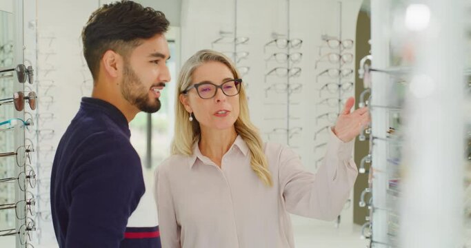 Female optician or optometrist helping a customer to choose glasses on the shelf in her shop. Friendly mature woman and owner helping a man to choose and try on spectacles in her optical store