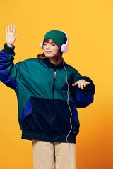a happy, joyful woman in a green jacket and hat enjoys music while standing in pink headphones on a yellow background dancing. Vertical photography