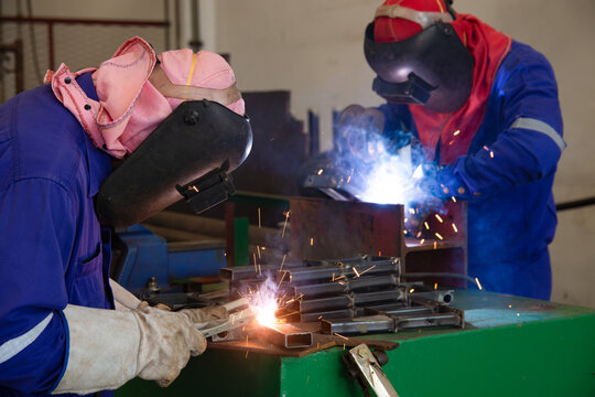 Two welder are assembling the workpiece by process shielded metal arc welding (SMAW). Welder in blue uniform, leather gloves, welding mask. The workers are testing a welding in the workshop. - Powered by Adobe
