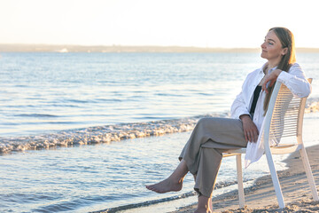 A beautiful woman sits on a chair near the sea.