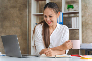 Fototapeta na wymiar Attractive Asian woman working with laptop, holding coffee cup at home office.